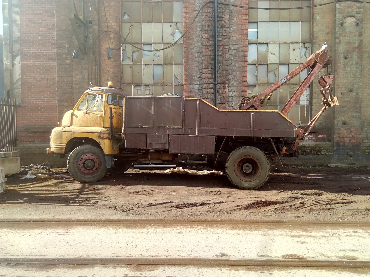 A Bedford RL truck parked in front of a very dilapidated
industrial building with most of the windows smashed out and boarded
from the inside. The truck has a faded yellow cab, and the body is
painted a dark chocolate brown.