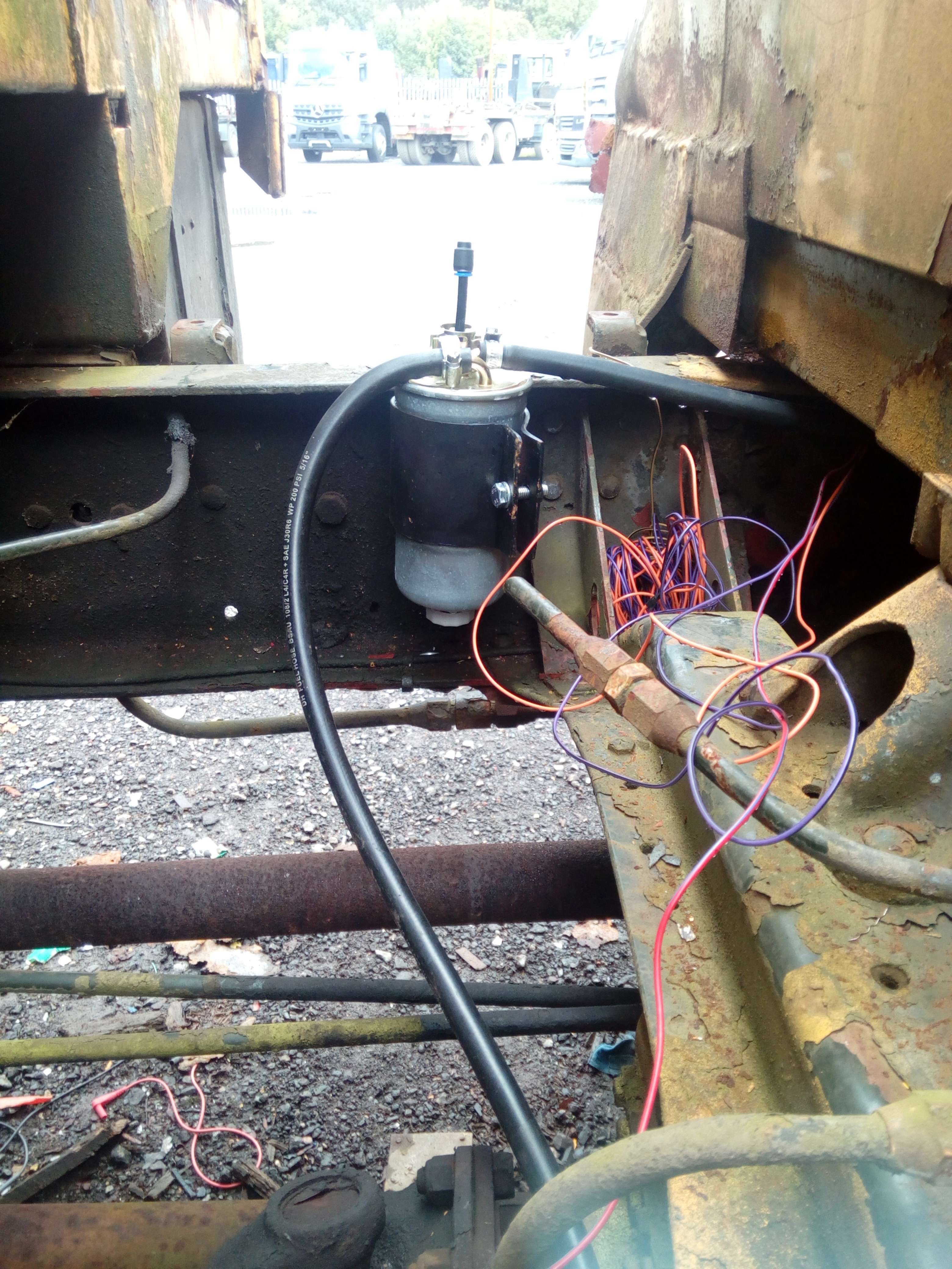 Photograph of the bracket bolted to the inside of the truck
shassis, with the filter stuffed into it and fuel lines
connected. Sticking out of the top of the fuel filter is a short
length of plastic pipe with a blanking cap on it. Piled up in the
chassis next to the filter is a large bundle of coloured wire.
