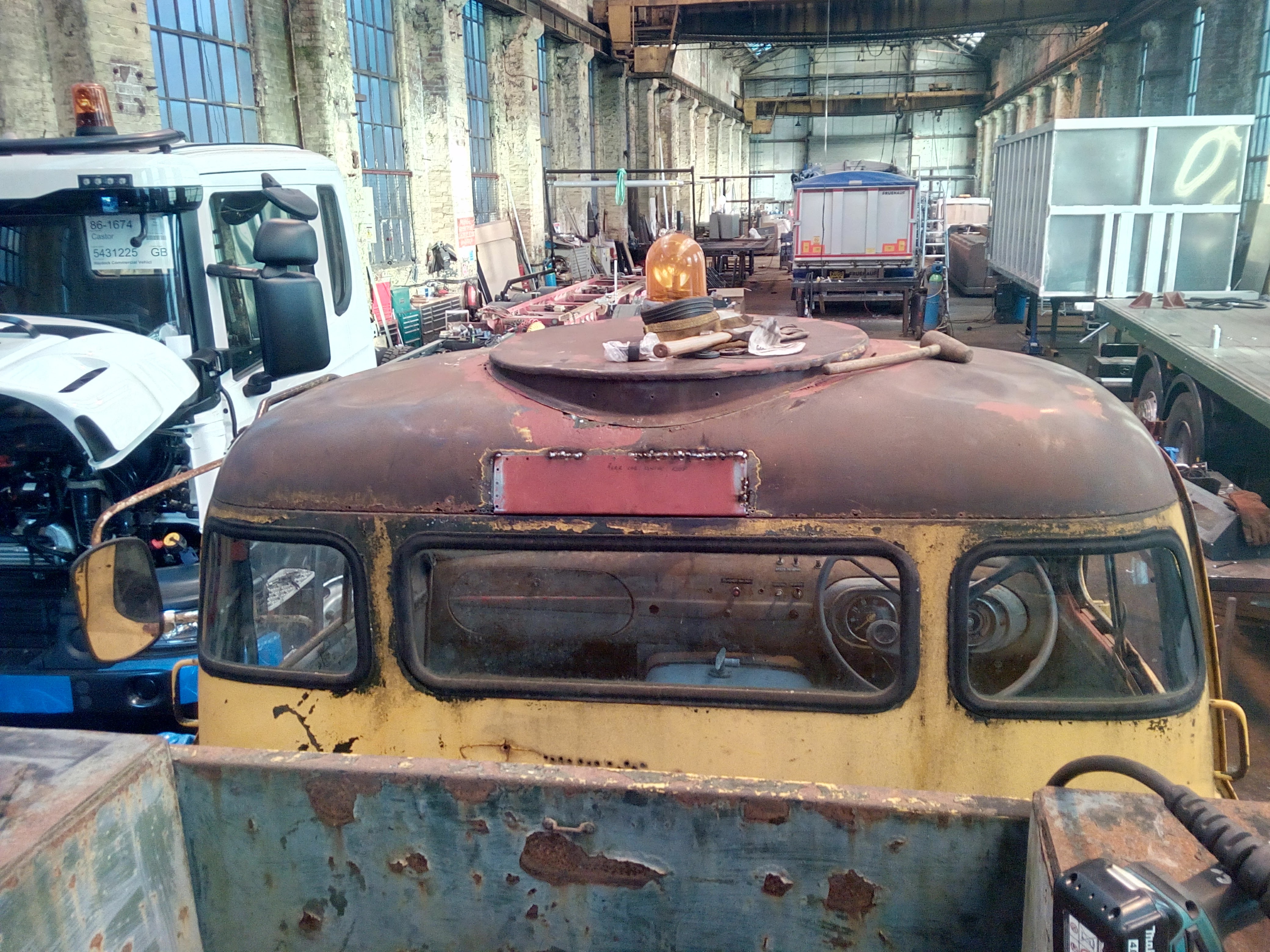 Photograph of the back of the truck cab, with the truck now
parked inside an old workshop building; visible all around the truck
are truck bodies in the process of being built, and a modern tipper
truck chassis. Overhead are two very large cranes running on tracks.