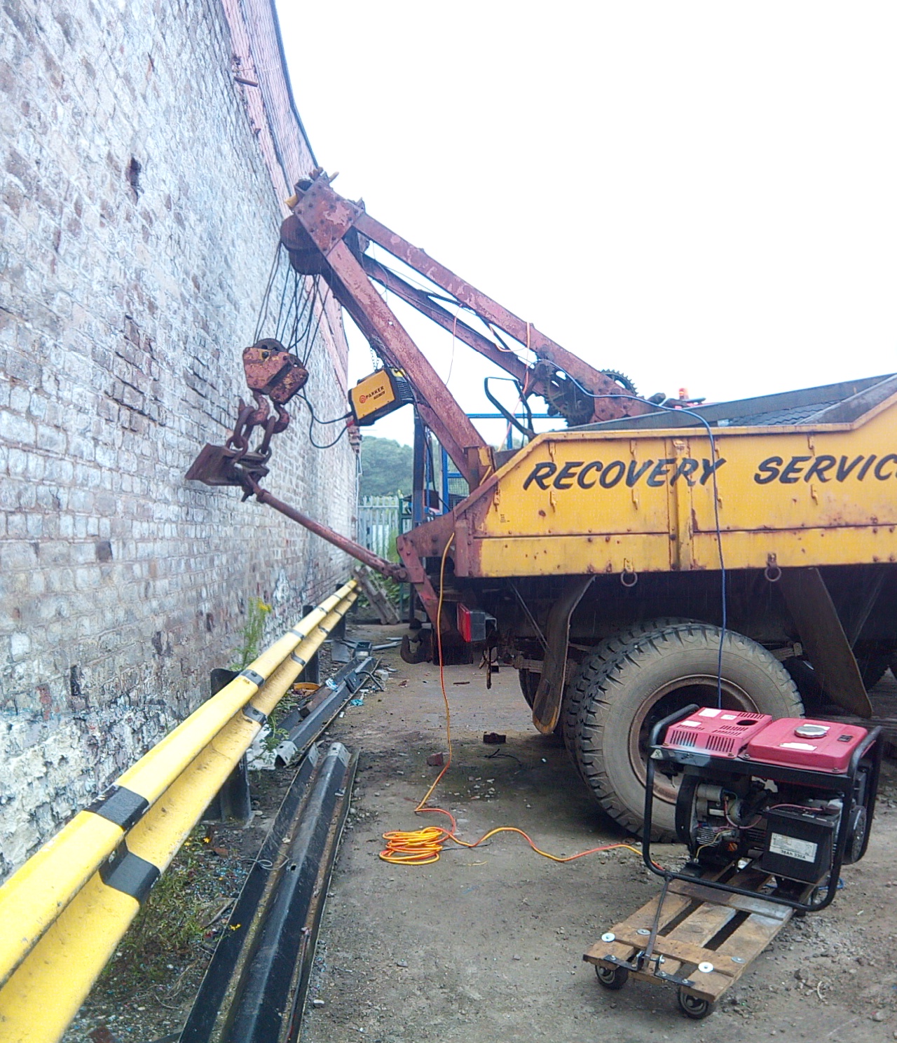 The truck backed up to a wall, with the recovery boom almost
touching said wall. A generator is set up in the foreground, and a
small stick welder is dangling from the boom by a length of chain.
