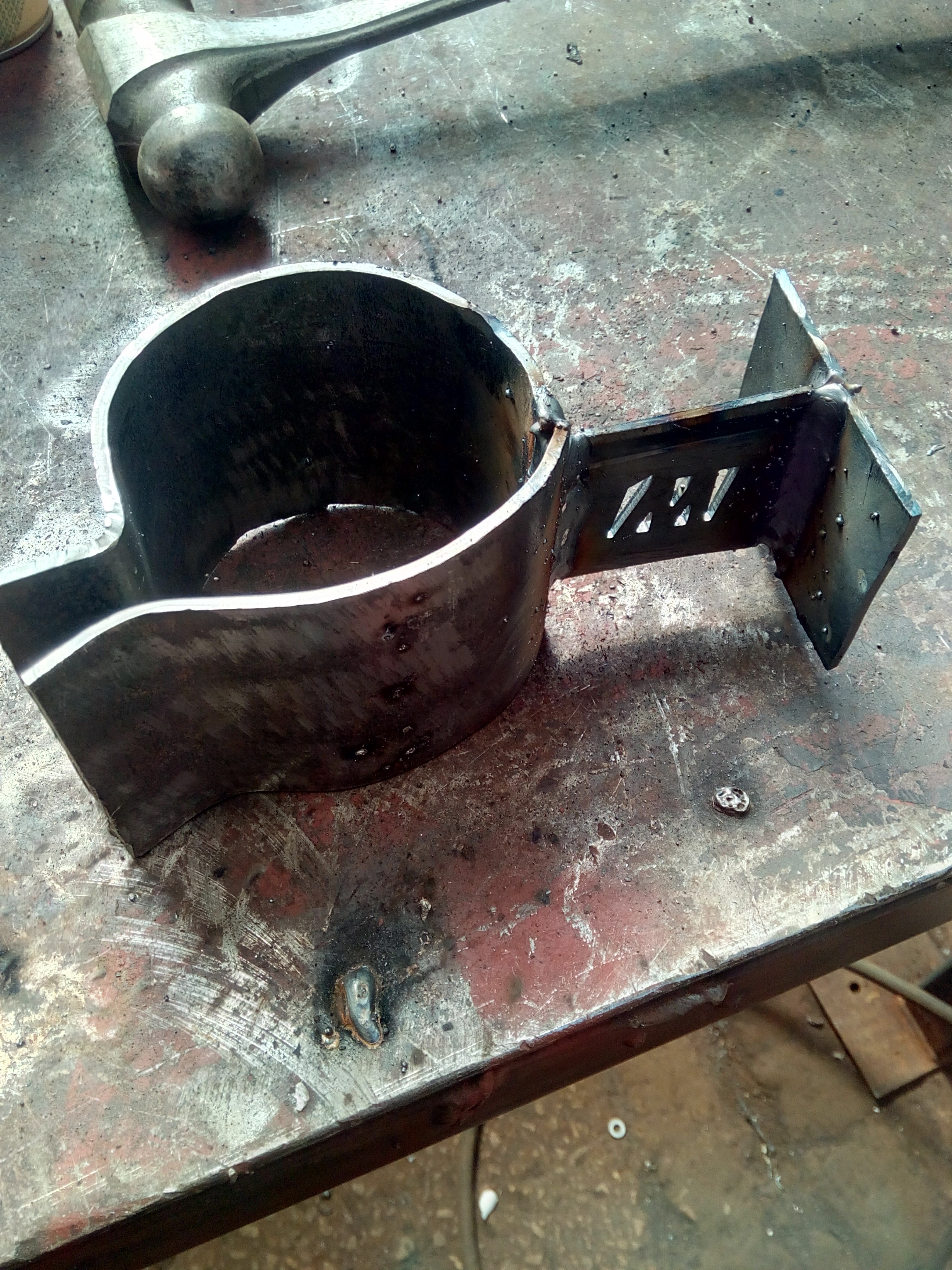 The same freshly-welded bracket, now sitting in a different
orientation on the welding table. The M logo, clamping bolt, and
several large globs of welding spatter, are clearly visible.