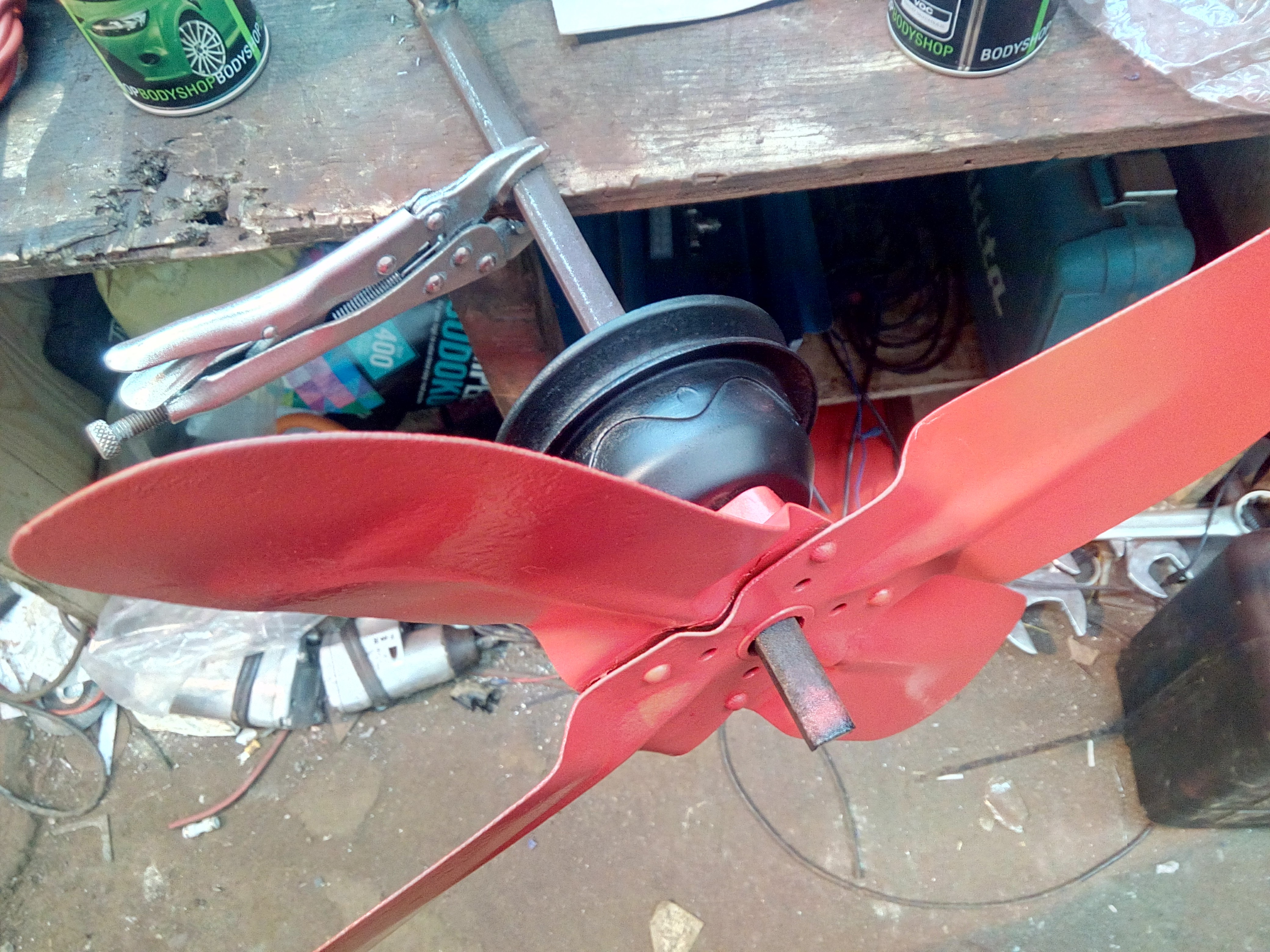 Photograph of the cooling fan and water pump pulley hanging off a
piece of metal rod clipped to the workbench. The fan is painted bright
red, and the pulley has been painted matte black.