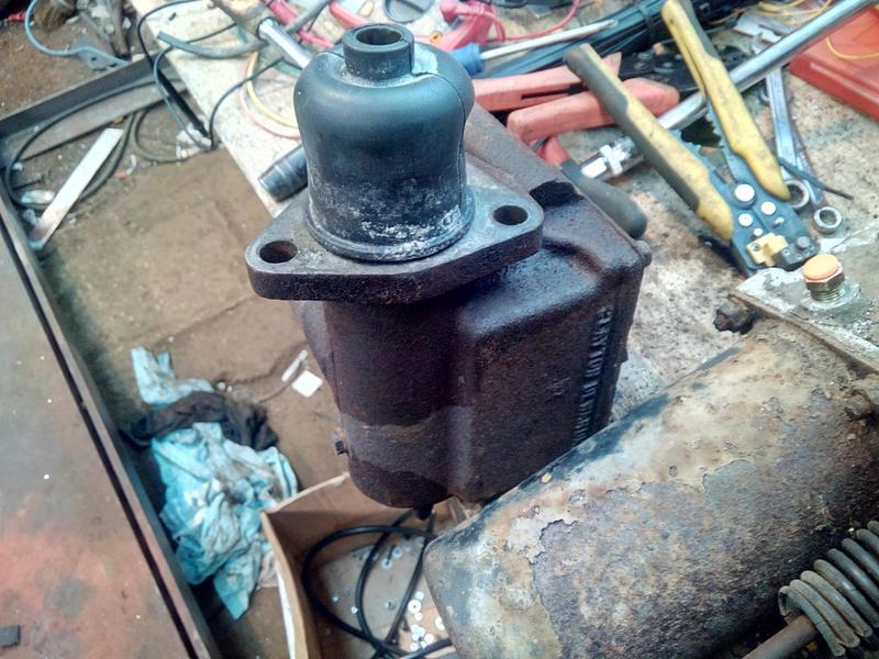 The very rusty brake master cylinder sat on its end on a cluttered work-bench. The rubber boot, meant to protect the open end of the cylinder, is pointed upwards.