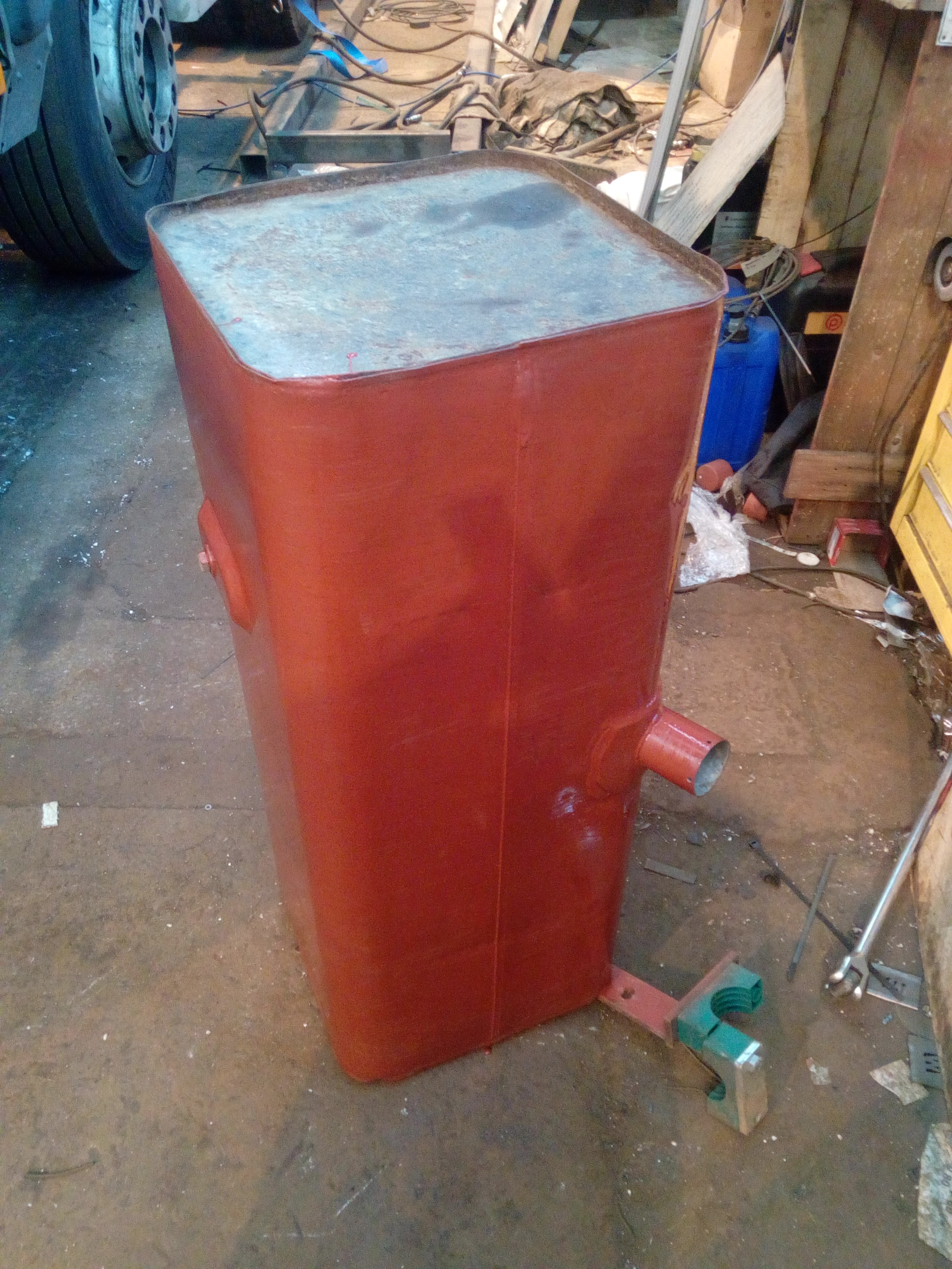A large rectangular fuel tank, standing on end in a cluttered
workshop, almost all painted with a reddish-brown primer paint. The
top-most surface, one of the end plates, has not yet been cleaned and
painted.