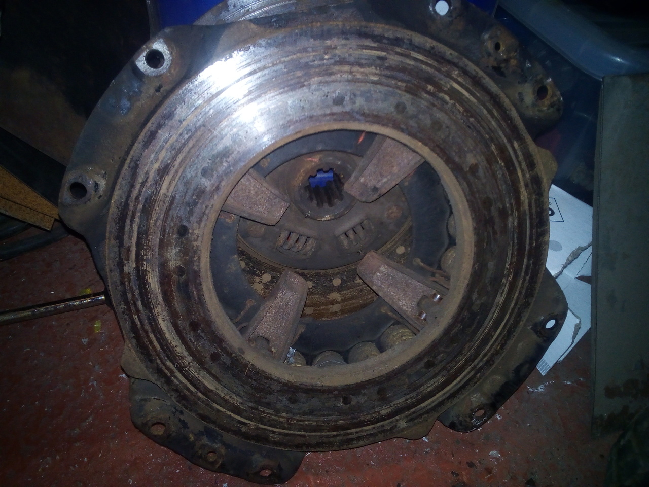 A clutch pressure plate leant up against a plastic box. The surface is very dark and corroded-looking, with shadow-imprints of the rivets of the friction plate still on it.