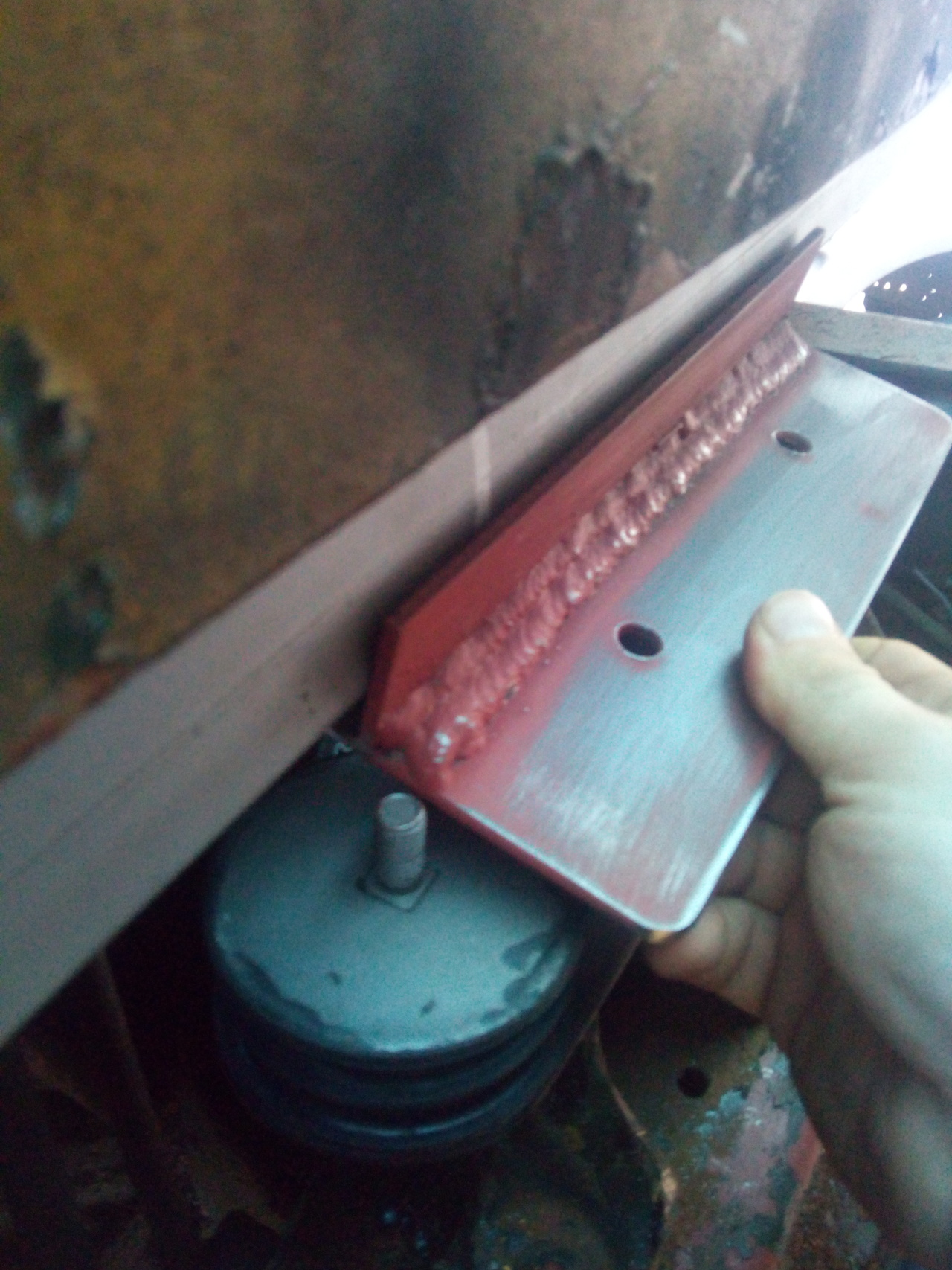 A hand holding up a bracket to the new rear cab cross-member. The
bracket has two holes drilled in it, for the top studs of the rubber
mounts, and a reinforcing tab that will help spread the load along the
crossmember. The reinforcing tab is in the wrong place and is
preventing the bracket from fitting.