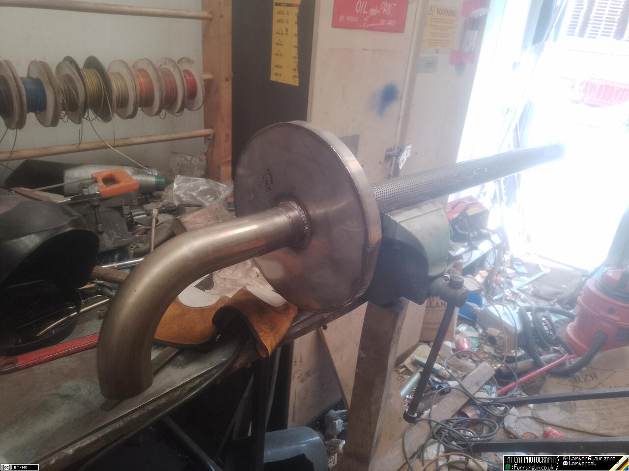 a large-radius 90 degree bend attached to some perforated tube, and stuck through the centre of the metal disk from previously; all of which is sitting on top of the vice