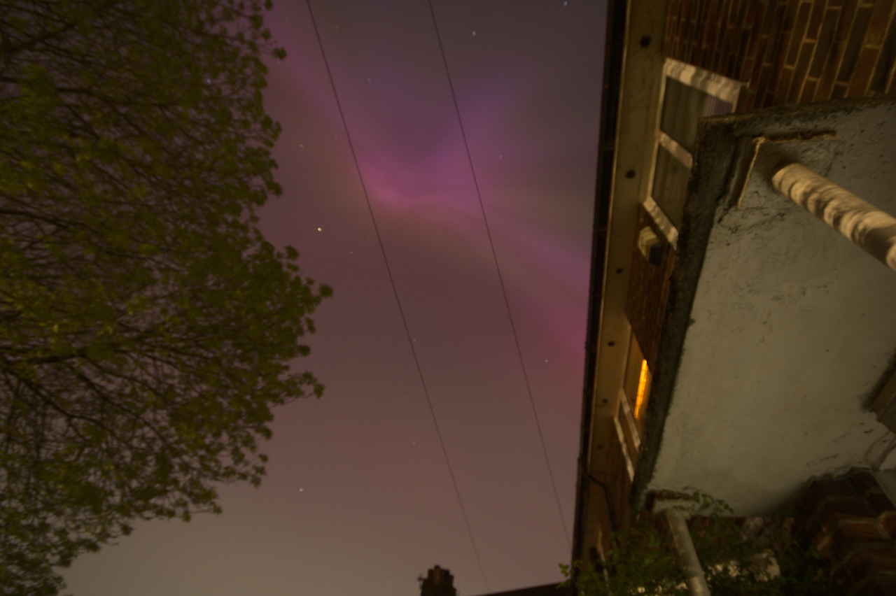 A long exposure photograph looking directly up in front of a house, with a pinkish-purple glow of the aurora spreading across the sky.