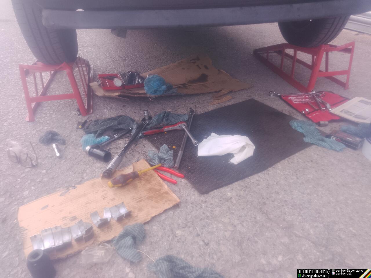 Looking at the floor in front of the car, which is up on ramps; there is some oily cardboard, a rubber mat, and several oily tools, as well as the original main and rod bearings