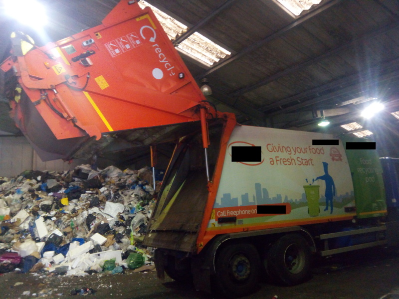 Garbage truck parked in front of a pile of garbage, with the back of the truck hinged open